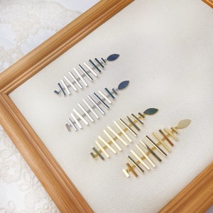 Peut inclure: Trois paires de boucles d'oreilles pendantes avec un design géométrique. Les boucles d'oreilles sont en métal argenté et doré et présentent une série de barres verticales avec un charme en forme de feuille au bas.