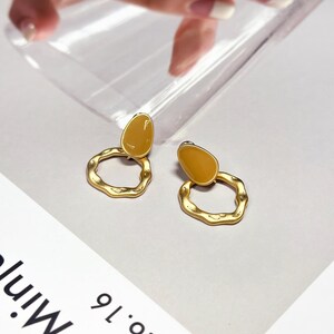May include: A pair of gold-coloured earrings. Each earring has a teardrop-shaped, mustard-yellow accent above a textured, irregular circle. The earrings are displayed on a white surface.