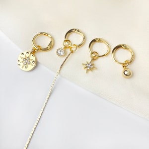 Peut inclure: Quatre boucles d'oreilles créoles dorées avec différents pendentifs. Un pendentif est un disque rond avec un motif en étoile, un autre est une étoile, un autre est une petite pierre ronde et le dernier est une petite boule ronde.