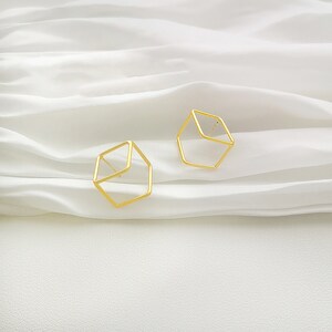 Puede incluir: Un par de pendientes de cubo geométrico de color dorado. Los pendientes están hechos de un alambre metálico fino y tienen un diseño simple y moderno.