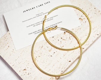 May include: Two large gold hoop earrings on a white textured surface. The earrings are made of a thick, round wire. A card with jewelry care tips is visible in the background.