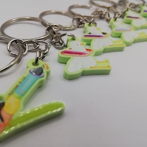 Könnte beinhalten: Schlüsselanhänger mit einem grünen und weißen Cartoon-Vogel-Design mit einem farbenfrohen Accessoire zum Thema Regenbogen. Die Schlüsselanhänger haben einen silbernen Metallring. Das Bild zeigt auch einen Schlüsselanhänger mit einem farbenfrohen Pinsel-Design.