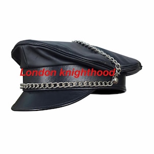 Può includere: Cappello in pelle nera con un dettaglio a catena argentata. La catena è attaccata alla parte superiore del cappello e scende lungo il lato.