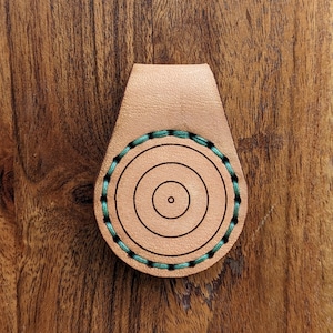 May include: A tan leather item with a stitched circular design. The circular design has concentric black lines and teal stitching around the perimeter. The item has a rectangular top and a circular bottom.