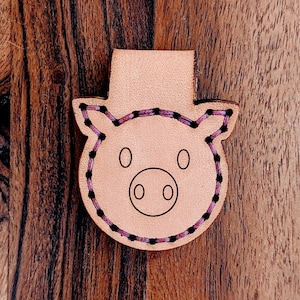 Puede incluir: Un marcapáginas de cuero marrón claro en forma de cerdo con costuras rosas alrededor de los bordes. El cerdo tiene ojos negros y un hocico.