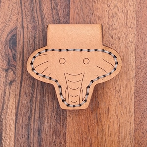Puede incluir: Un marcapáginas de cuero marrón claro en forma de elefante con un contorno cosido y dos pequeños círculos para los ojos.