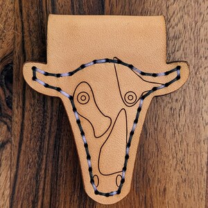 Puede incluir: Un llavero de cuero marrón con un diseño de cabeza de vaca cosido. Las costuras son negras y moradas.