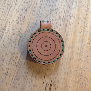 May include: A brown leather target with a green stitched border. The target has concentric circles.