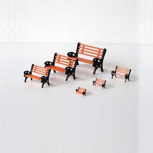May include: A collection of miniature park benches. The benches feature orange seats and black frames with decorative armrests. The benches are arranged in different sizes, suggesting a park scene. The smallest bench is approximately 2.5 cm long.