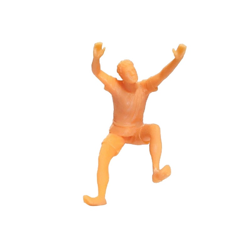May include: A small, orange plastic figurine of a person with arms raised in the air. The figurine is wearing shorts and has a simple, cartoon-like design.