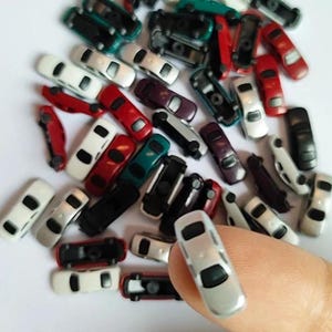 May include: A collection of miniature toy cars in various colors, including red, white, black, and silver. The cars are small, with visible details like windows and wheels. One silver car is held by a finger, showcasing the size of the toys.