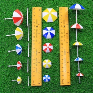 Pode incluir: Um conjunto de 10 mini guarda-sóis de praia em várias cores, incluindo vermelho, branco, azul e amarelo. Os guarda-sóis são feitos de plástico e têm um poste de plástico branco. Os guarda-sóis estão dispostos em duas filas sobre uma superfície verde com uma régua para escala.
