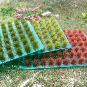 May include: Close-up of three rectangular model grass mats in shades of green, yellow-green, and red-brown. Each mat features dense, tufted grass-like fibers attached to a transparent green base. The mats are arranged on a surface with scattered green and brown ground cover.