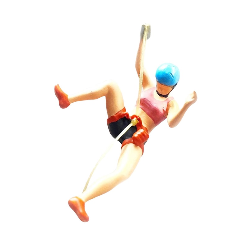 May include: A figurine of a person rock climbing, wearing a pink sports bra, black shorts, and a blue helmet. The figurine is attached to a white rope.
