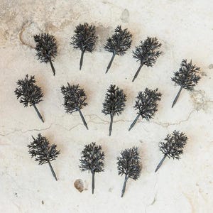 May include: A collection of 15 black miniature trees, arranged in a circular pattern on a light grey surface. The trees are made of a dark material and have a realistic appearance.