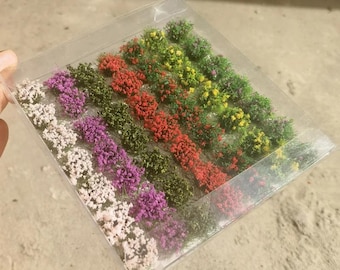 Könnte beinhalten: Ein durchsichtiger Kunststoffbehälter mit einer Sammlung von künstlichen Miniatur-Blumenbüscheln in verschiedenen Farben wie Weiß, Lila, Grün, Rot und Gelb. Diese kleinen, strukturierten Blumenbüschel sind in ordentlichen Reihen in der Box angeordnet.