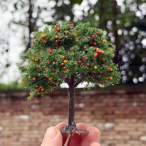May include: A small model tree with a brown trunk, green leaves, and tiny red and orange fruits. The tree is held in a hand, with a blurred backdrop of foliage and a brick wall.