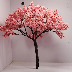 Puede incluir: Un árbol artificial de cerezo en flor rosa con un tronco y ramas de color marrón oscuro. El árbol está en una habitación blanca con una pared y un suelo blancos.