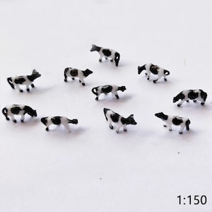 Op de afbeelding: Miniatuur zwarte en witte koeien verspreid over een witte ondergrond. De kleine beeldjes zijn gedetailleerd met geschilderde vlekken en staan in verschillende houdingen. De afbeelding bevat de tekst "1:150" rechtsonder.