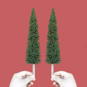 以下が含まれることがあります： 人の手に持たれた、茶色の土台が付いた緑色のクリスマスツリーが2本