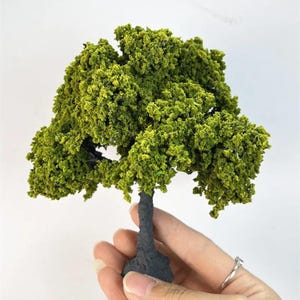 May include: A small model tree featuring a dense, bright green foliage and a dark grey trunk. This miniature tree is suitable for model railways, dioramas, or craft projects. The tree is held in a hand against a white backdrop.
