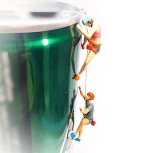 May include: Two miniature figures are climbing a green can. The figures are wearing climbing gear and are attached to ropes. The can is a popular brand of soda.