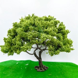 May include: A miniature tree model with a dark brown trunk and branches, and a full, bright green canopy. The tree sits on a patch of green, artificial grass against a white background. Ideal for model train sets or dioramas.