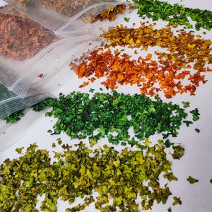May include: An overhead shot displays several small plastic bags filled with dried, colorful plant matter. The contents are spilling out onto a white surface, creating rows of orange, green, yellow, and brown hues. The dried material appears to be small, leaf-like pieces.