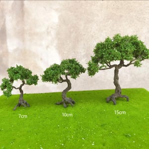 Puede incluir: Tres árboles artificiales en miniatura con follaje verde y troncos marrones sobre una superficie verde. Los árboles miden 7 cm, 10 cm y 15 cm de alto.