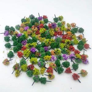 Puede incluir: Una colección de árboles modelo en miniatura en varios colores, incluyendo verde, rojo, amarillo y morado. Cada árbol tiene un pequeño tallo oscuro. Estos pequeños árboles se utilizan probablemente para maquetas de trenes, dioramas u otros proyectos de manualidades.