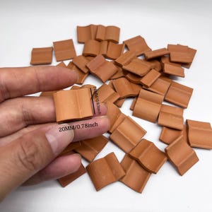 May include: A collection of small, curved, terracotta-coloured clay roof tiles. One tile is held up to show dimensions of 16mm by 20mm. The tiles have a distinct arched shape and a matte, earthy finish, suitable for miniature construction projects.