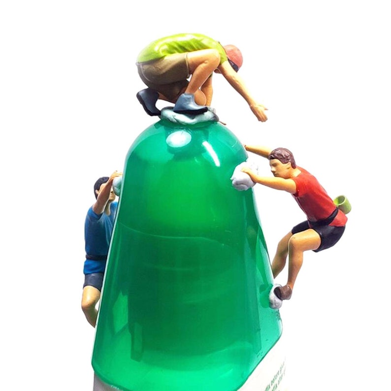 May include: Three miniature figures are climbing a green plastic bottle. The figures are wearing colourful clothing and are in various poses. The bottle is clear and has a textured surface.