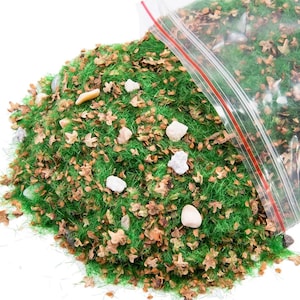 Op de afbeelding: Een verzameling modelspoorweglandschapsmaterialen, waaronder groen statisch gras, kleine bruine bladeren en witte en beige stenen. De materialen komen uit een doorzichtige plastic zak met een rode ritssluiting.