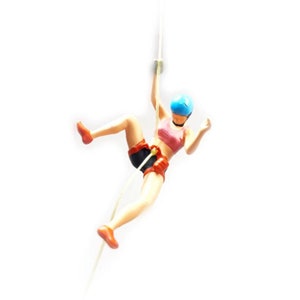 May include: A figurine of a person abseiling down a rope, wearing a blue helmet, pink sports bra, black shorts, and red climbing gear.