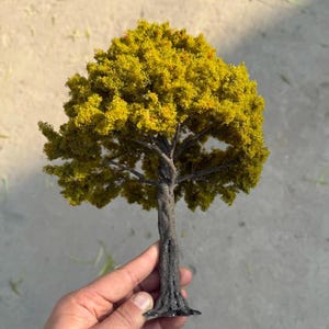 Könnte beinhalten: Ein Miniaturmodell eines Baumes mit einem strukturierten, dunkelbraunen, gedrehten Stamm und einer dichten, abgerundeten Baumkrone, die mit leuchtend gelb-grünem Laub bedeckt ist. Die Struktur ahmt einen Laubbaum nach, mit feinen Ästen unter dem Laub.