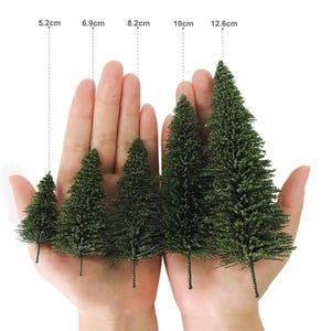 May include: Five artificial pine trees of varying sizes, ranging from 5.2 cm to 12.6 cm tall, are displayed in a hand. The trees are green and have a fluffy texture.