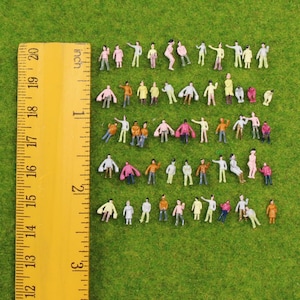 Può includere: Una collezione di figurine in plastica in miniatura che rappresentano persone in varie pose e abiti colorati, disposte in file accanto a un righello giallo che mostra una scala di 7,62 cm, fornendo un chiaro riferimento visivo per le dimensioni.