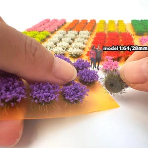 May include: A variety of colours of artificial grass tufts, including purple, pink, white, yellow, and green. The tufts are arranged in rows on a sheet of orange paper. The text "model 1:64/28mm" is visible in the image.