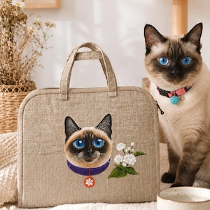 Puede incluir: Un bolso rectangular de lino beige con un bordado de un gato siamés de ojos azules brillantes, collar morado y un pequeño dije rojo, acompañado de flores de jazmín blancas en el panel frontal y dos asas superiores para su transporte.