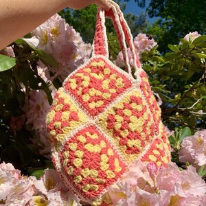 May include: A crocheted tote bag with a yellow and pink granny square pattern. The bag has two straps and is made with a white yarn.
