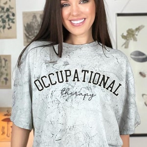 May include: A light grey t-shirt with the words "OCCUPATIONAL therapy" printed in black. The shirt has a textured, distressed appearance. The person is wearing a white watch.