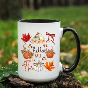 May include: White ceramic mug with a black rim and handle. The mug features a fall-themed design with pumpkins, leaves, and the text "hello fall".