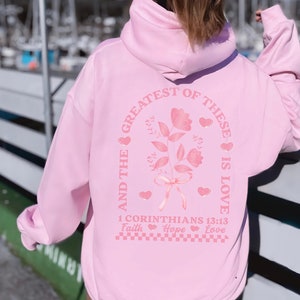May include: Pink hoodie with a floral design and the text "And the greatest of these is love 1 Corinthians 13:13 Faith Hope Love" printed on the back.