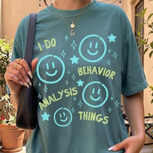 Könnte beinhalten: Ein kurzärmeliges T-Shirt in Blaugrün mit einem Grafikdruck aus hellblauen Smileys, Sternen und Funken. Der Text lautet I DO BEHAVIOR ANALYSIS THINGS in hellgrüner und hellblauer Schrift. Das Shirt hat einen Rundhalsausschnitt.