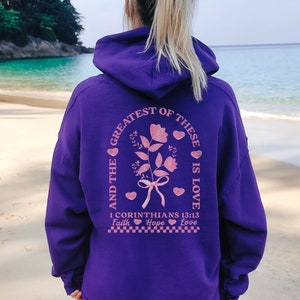 May include: Purple hoodie with a pink graphic on the back that says "And the greatest of these is love 1 Corinthians 13:13 Faith Hope Love" with a floral design and hearts.