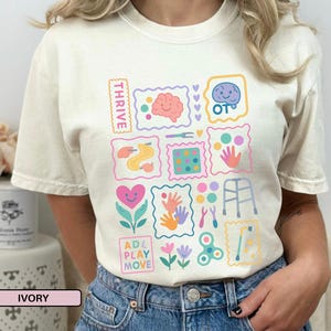 Könnte beinhalten: Elfenbeinfarbenes T-Shirt mit einem farbenfrohen Grafikdesign. Das Design umfasst verschiedene Illustrationen wie ein Gehirn, Hände und Blumen, zusammen mit den Wörtern "THRIVE" und "ADL PLAY MOVE". Das Shirt ist aus weichem Material.