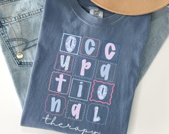 Op de afbeelding: Een blauw Comfort Colors T-shirt met de woorden "Occupational Therapy" in een kleurrijk, omlijnd ontwerp. Het shirt ligt op een spijkerbroek en een bruine hoed.