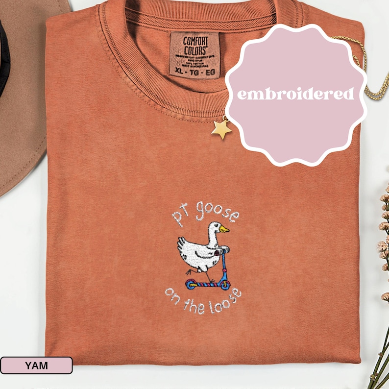 May include: A terracotta-colored t-shirt with an embroidered design of a white goose riding a blue scooter. The text "pt goose on the loose" is embroidered below the goose. The shirt has a Comfort Colors tag.