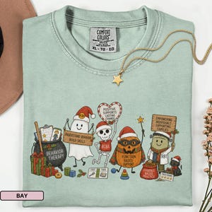 Op de afbeelding: Een lichtgroen t-shirt met een afbeelding van een geest, skelet, pompoen en het monster van Frankenstein met kerstmutsen. De tekst bevat "BEHAVIOR THERAPY," "UNDERSTAND BEHAVIOR BUILD SKILLS," "POSITIVE SUPPORTS LASTING CHANGE," en "EMPOWERING INDIVIDUALS SUPPORTING FAMILIES."