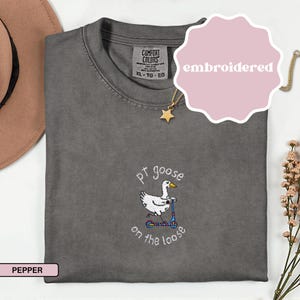 May include: Pepper-colored Comfort Colors t-shirt with an embroidered design. The design features a white goose riding a blue scooter, with the text "pt goose on the loose". A pink sticker with the word "embroidered" is on the top right.